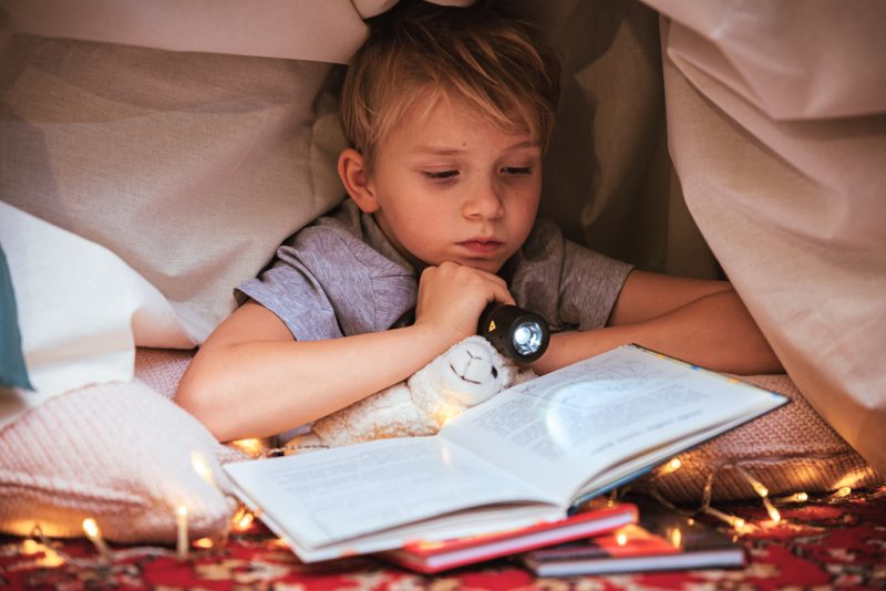 Pieni poika lukee kirjaa taskulampun valossa majassa.