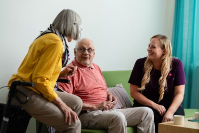Kaksi ikääntynyttä ja hoitaja keskustelevat hyväntuulisina sohvalla.