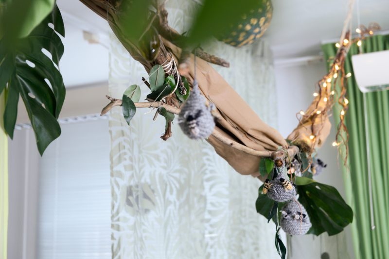 Katosta roikkumassa koriste, jossa ruskeaa kangasta, vihreitä viherkasvien lehtiä, valoja sekä kudottuja ampiaisenpesiä ampiaisineen.