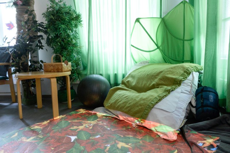 Vihreänsävyinen kuva, jossa seinustalla vihreiden verhojen edessä lattialla säkkituoli. Lisäksi huoneessa jumppapallo ja pieni pöytä. Lattialle heijastuu syksyn lehtiä eri väreissä.