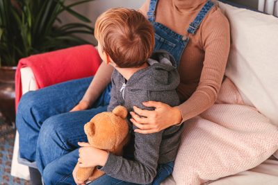 A child and an adult are sitting in a couch. A child is holding teddy bear in his lap. An adult folds child from his shoulders.