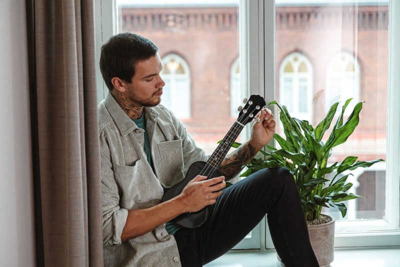 A man is sitting in front of a window and playing a guitar.