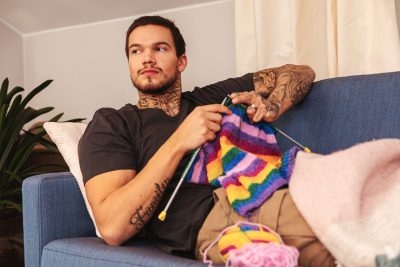 A man is sitting on a couch and knitting a colorful scarf.