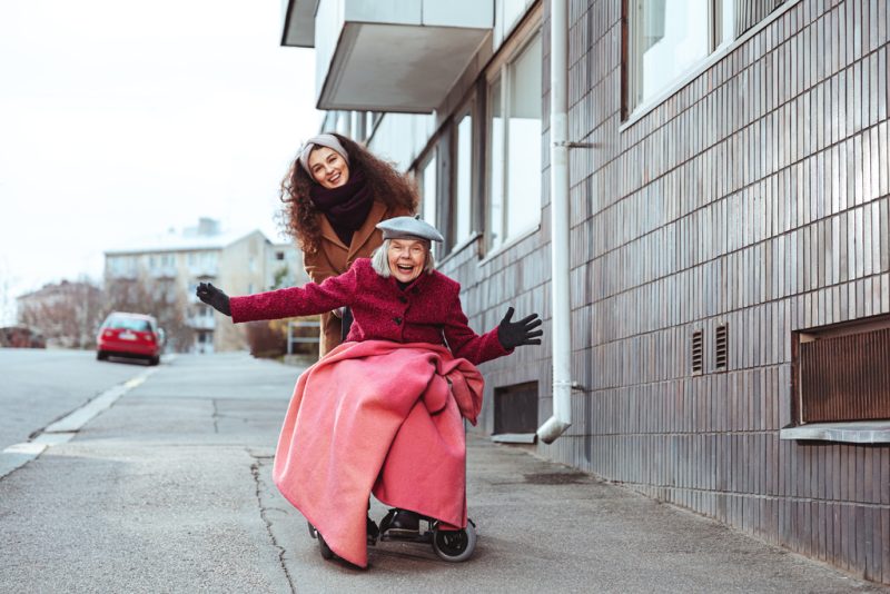 A woman in pushing an elderly woman in a wheelchair outside in the walkway.