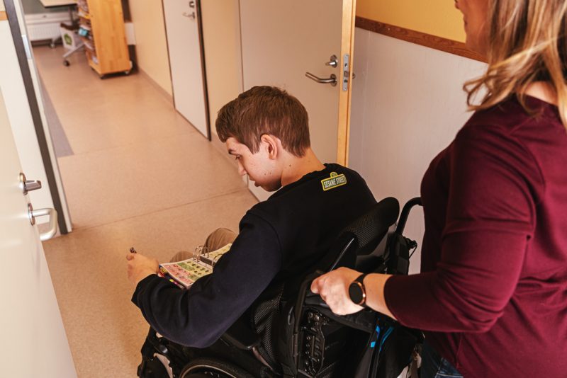 A person is pushing a young man in a wheelchair inside the corridor.