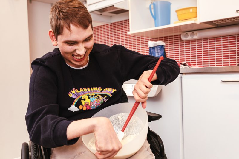 A person sitting in a wheelchair is whipping something in a bowl. He is holding the bowl and smiling.