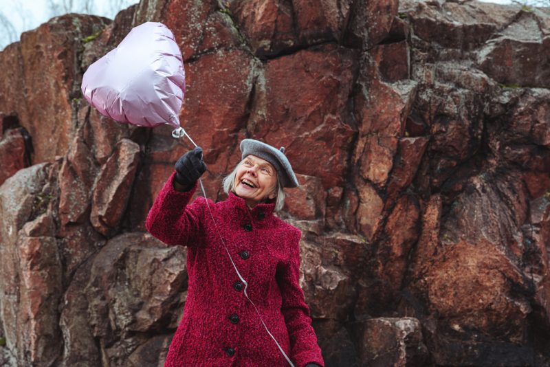 An elderly woman is holding a heart shaped ballon in her hands while standing outside in front of a big rock.