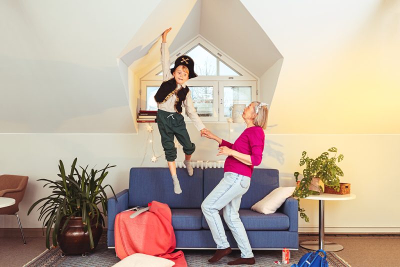 A child dressed up as a pirate is jumping high in the air from couch. An elderly woman is holding his hand and seeing him.