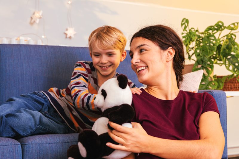 A child is sitting on a couch. A woman is sitting in a ground in front of child, holding a panda teddy bear in her hands. Both are smiling.