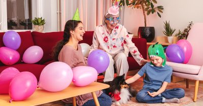 Lapsi ja nainen istuvat lattialla koiran vieressä. Vanhempi nainen istuu sohvalla. Kuvassa on juhlatunnelmaa - ilmapalloja, naamioita ja muita koristeita.