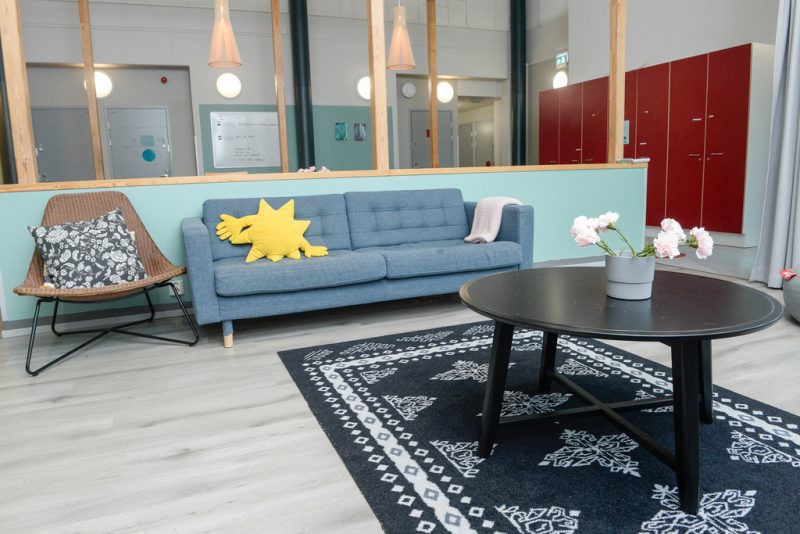 Oleskelutilassa sinisen sävyinen sohva, ruskea nojatuoli ja mustan koristeellisen maton päällä musta sohvapöytä, jonka päällä kukkia maljakossa. Taustalla näkyy toimipisteen muita tiloja ja käytävää.