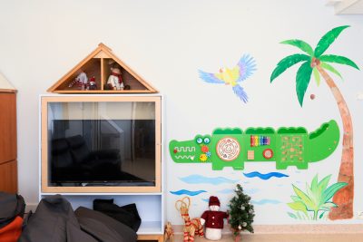 Tv-tason ja television vieressä seinään kiinnitetty krokotiilin muotoinen aktiviteettiseinä, jossa erivärisiä aktivoivia tehtäviä. Seinällä myös palmu ja papukaija maalaukset.