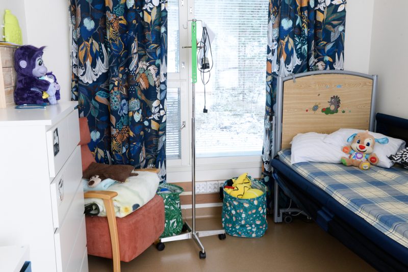 Lapsen huone, jossa sänky, nojatuoli ja korkeahko valkoinen hyllykkö. Ikkunoissa tummansiniset koristeelliset verhot. Lattialla laatikoissa pehmoleluja.