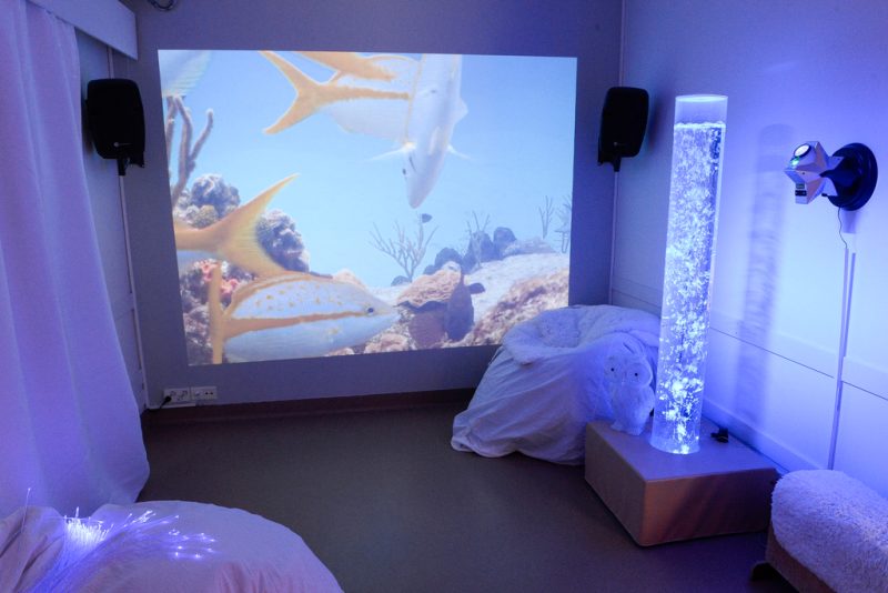 Sinisensävyinen aistihuone, jossa valkoisia säkkituolia. Seinälle heijastettu akvaarionäkymää.