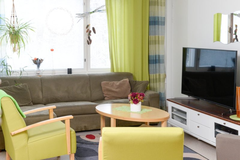 Oleskelutilassa tummanruskea sohva ja kaksi vaaleanvihreää nojatuolia. Keskellä huonetta sohvapöytä. Seinustalla matala tv-taso ja taulutelevisio.