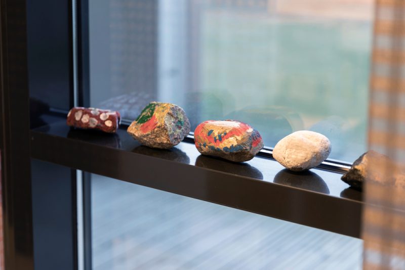 Ikkunalaudalla mustan kaiteen päällä viisi eri värein maalattua erimuotoista kiveä.