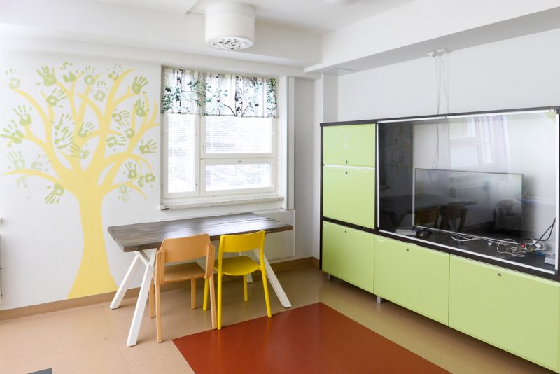 Oleskelutilassa ikkunan edessä pöytä ja kaksi tuolia, yhdellä seinustalla iso vihreänsävyinen televisiohylly. Seinään on maalattu keltainen puunrunko ja vihreällä käsien jälkiä ikään kuin lehdiksi.