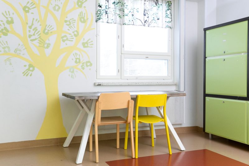Ikkunan edessä pieni pöytä, jonka äärellä kaksi tuolia. Seinään on maalattu keltainen puunrunko ja vihreällä käsien jälkiä ikään kuin lehdiksi. Toisella seinustalla vihreäovinen kaappi.