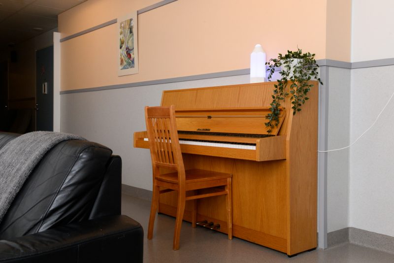 Harmaan ja beigen sävyisen seinän edustalla puun värinen piano ja tuoli. Pianon päällä viherkasvi.