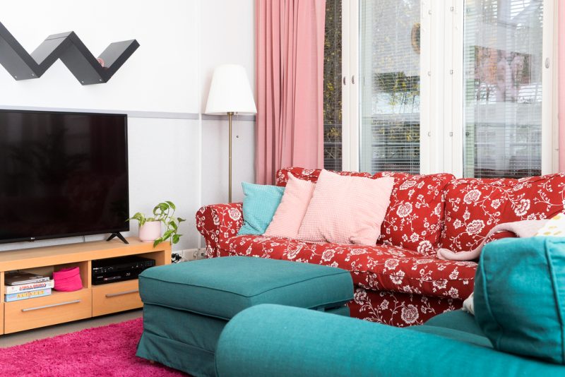 Kuva sohvaryhmästä ja televisiosta oleskelutilassa. Sohvan takana isot ikkunat metsäiseen näkymään. Huonekalujen sisustuksessa on käytetty raikkaasti eri värejä: turkoosia, punaista, vaaleanpunaista ja valkoista.