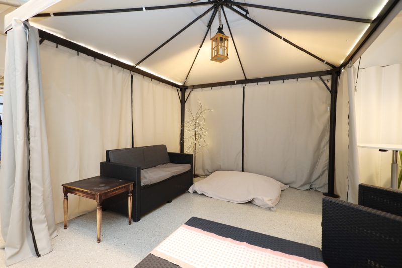 Vaalea telttakatos, jonka sisällä mustia sohvia ja tuoleja, valkoinen säkkituoli sekä tunnelmallisia valoja.