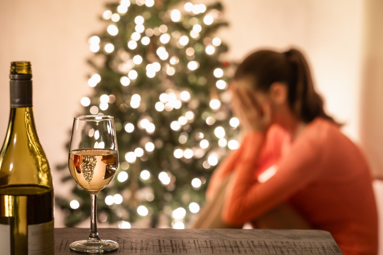 Etualalla viinipullo ja -lasi, taustalla valaistu joulukuusi ja sen edessä istumassa nainen, joka nojaa kasvoja käsiinsä.