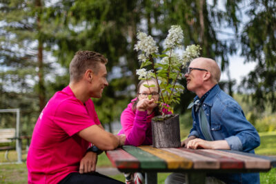 Kolme henkilöä istuu ulkona pöydän ympärillä. Pöydällä on puisessa maljakossa vaaleita kukkia. Ihmiset katsovat ja tuoksuttelevat kukkia.