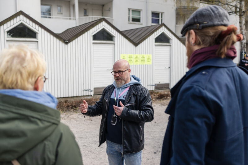 Kolme ihmistä keskustelemassa ulkona, kaksi selin kameraan, yksi kasvot kameraan.