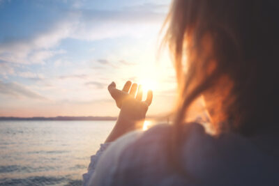 Nainen kurkottaa kädellään aurinkoa kohden veden äärellä.