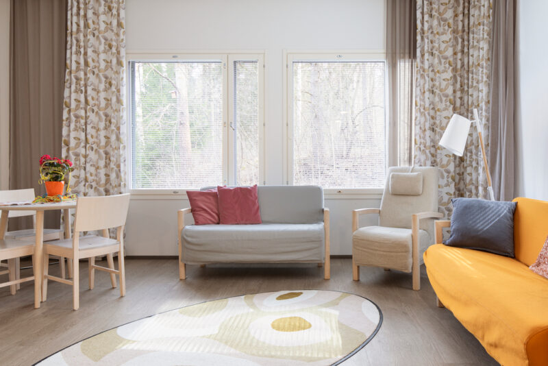 Oleskeluhuoneessa keltainen ja harmaa sohva, nojatuoli sekä pöytä ja muutama tuoli sen ympärillä. Takana seinällä kaksi ikkunaa, joista metsänäkymä.