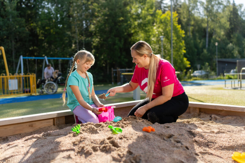 Lapsi ja Rinnekotien paitaan pukeutunut hoitaja hiekkalaatikolla, taustalla lapsi pyörätuolissa hoitajansa kanssa.