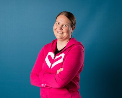Pinkkiin Rinnekotien brändihuppariin pukeutunut nainen katsoo kameraan hymyillen.