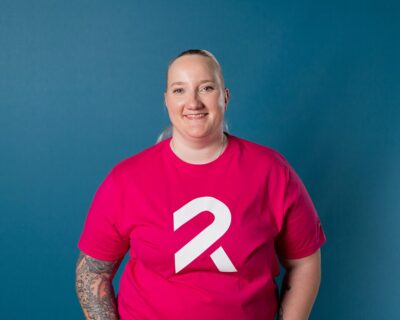 Pinkkiin Rinnekotien t-paitaan pukeutunut nainen katsoo kameraan hymyillen.