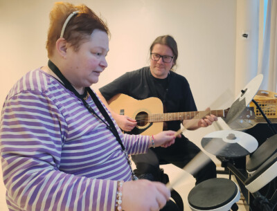 Kaksi henkilöä istuu musisoimassa yhdessä. Toinen henkilö soittaa rumpuja ja toinen akustista kitaraa.