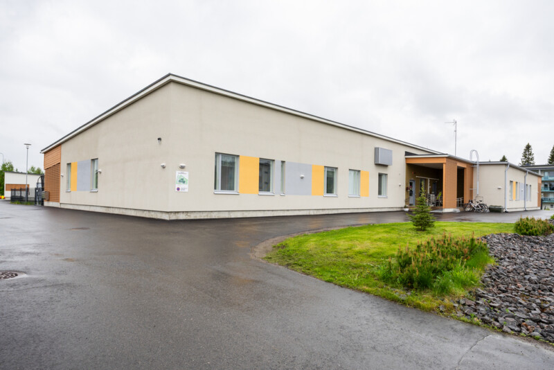 Yksikerroksisen rakennuksen julkisivu yhdestä kulmasta kuvattuna. Rakennus on vaalea ja sen edustalla on asfaltoitua pihaa ja viheraluetta.