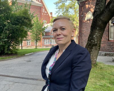 Vaalea nainen katsoo kameraan, taustalla punatiilinen rakennus ja puita.