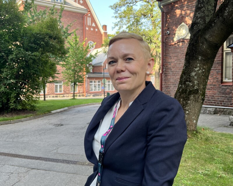 Vaalea nainen katsoo kameraan, taustalla punatiilinen rakennus ja puita.