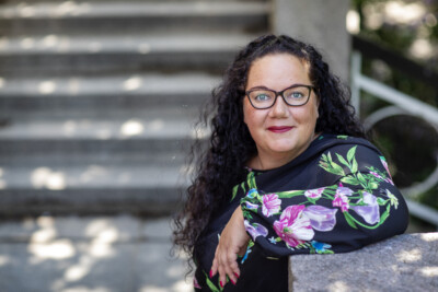 Nainen nojaa ulkona kiviseen tasoon ja katsoo hymyillen kameraan.