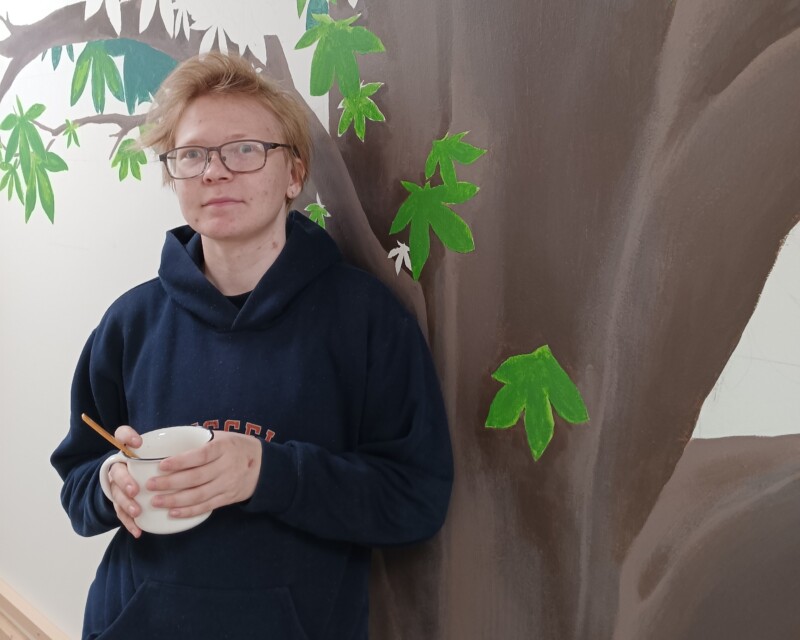 Nuori ihminen seisoo puuta esittävän maalauksen edessä kahvikuppi kädessään.
