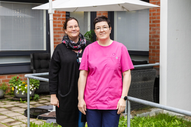 Kaksi henkilöä seisoo vieretysten kameraan katsoen ulkona punatiilisen rakennuksen edustalla.