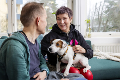 Nainen Rinnekodit-hupparissa pitää sylissään pientä valko-ruskeaa koiraa, ja juttelee vieressään istuvalle henkilölle, joka on selinkameraan.