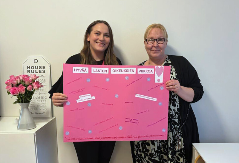 Kaksi henkilöä seisovat kasvot kameraan päin ja pitävät käsillään kiinni isosta vaaleanpunaisesta julisteesta, jossa on teksti Hyvää lasten oikeuksien viikkoa.