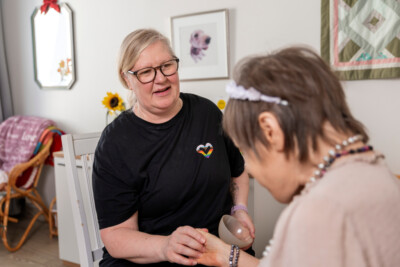 Mustaan t-paitaan pukeutunut nainen pitelee toista naista kädestä, nainen on selin kameraan.