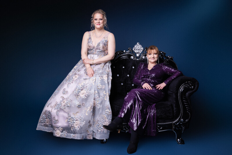 Kaksi naista juhlapuvuissa istuu mustassa isossa nojatuolissa.