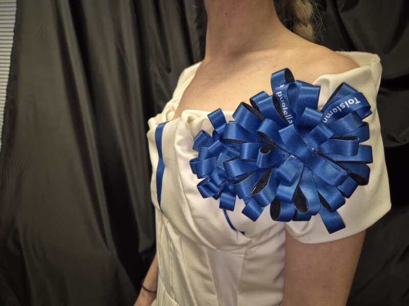 Valkoinen juhlapuku, jossa sininen ruusuke, jossa teksti Toistemme puolella.