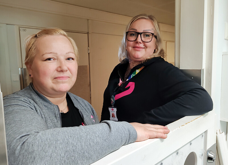 Kaksi henkilöä seisoo matalan oven takana ja katsovat kameraa kohti.