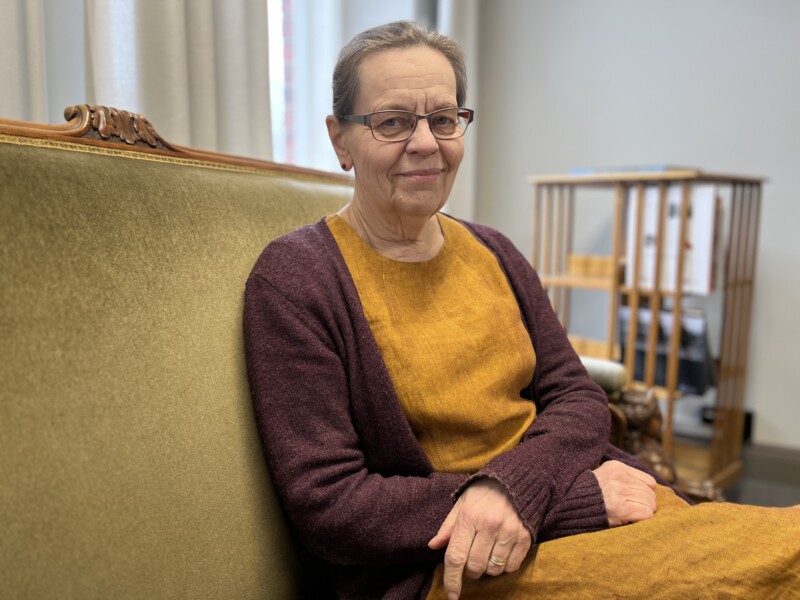 Nainen istuu kellertävässä nojatuolissa ja katsoo kameraan.