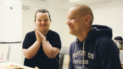 Kaksi henkilöä istuu pöydän ääressä ja toinen taputtaa käsiään yhteen.