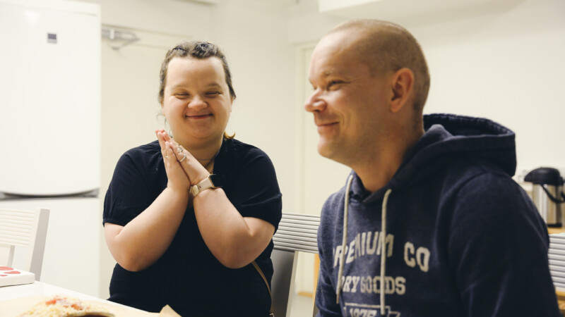 Kaksi henkilöä istuu pöydän ääressä ja toinen taputtaa käsiään yhteen.