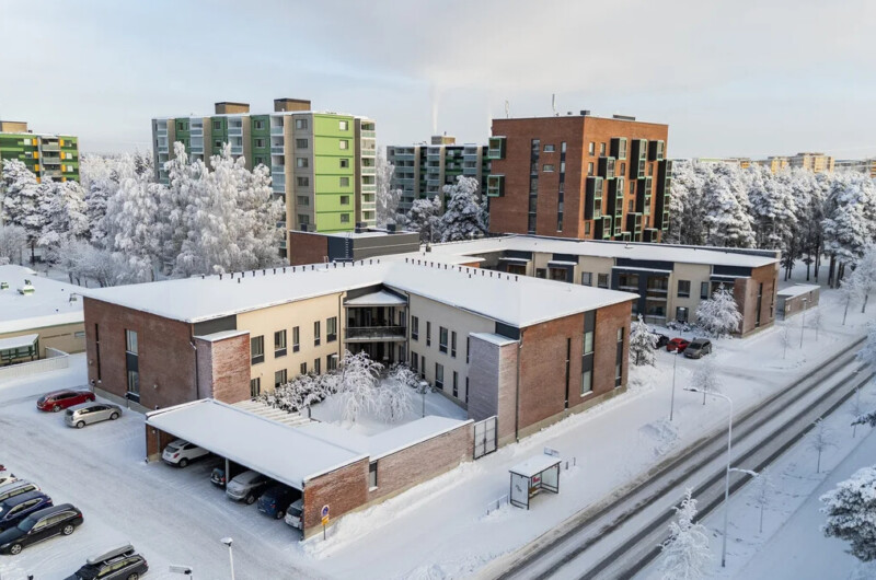 Miljöökuva, jonka keskiössä kaksikerroksinen, kulmikas rakennus. Taustalla näkyy korkeampia kerrostaloja. Maisema on luminen.